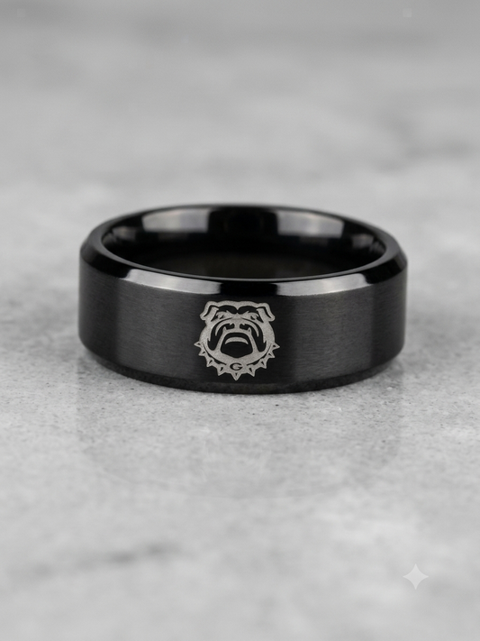 A black metallic band ring with a brushed finish and a silver bulldog emblem in the center, resting on a light gray stone surface.