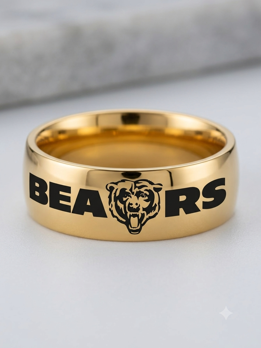 A polished gold band ring engraved with the word 'BEARS' and a bear head logo, shown on a light gray surface with a blurred gray background.