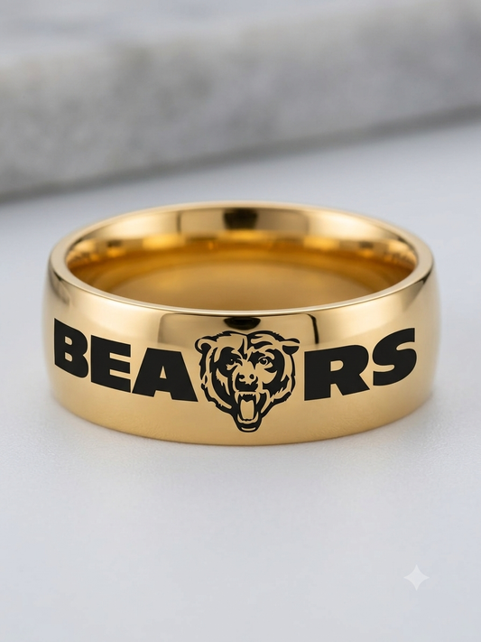 A polished gold band ring engraved with the word 'BEARS' and a bear head logo, shown on a light gray surface with a blurred gray background.